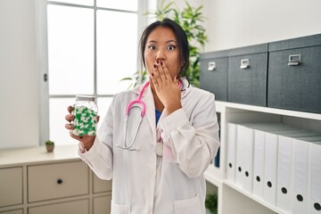 Young asian doctor woman holding pills covering mouth with hand, shocked and afraid for mistake....