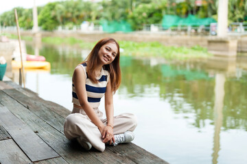 Fototapeta na wymiar Cute Asian girl with long hair sitting and relaxing in her free time on the pier By the river on weekends with smiling faces A rural canal with clear water