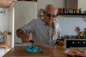 eighty-year-old man in the kitchen with a rag on his shoulder with a bread knife, cleaning the chopping wood with a wooden spoon.
