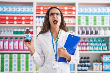 Young hispanic woman working at pharmacy drugstore holding syrup angry and mad screaming frustrated...