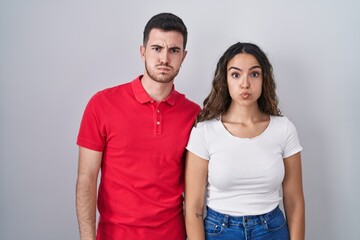 Young hispanic couple standing over isolated background puffing cheeks with funny face. mouth inflated with air, crazy expression.