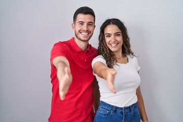 Young hispanic couple standing over isolated background smiling friendly offering handshake as greeting and welcoming. successful business.