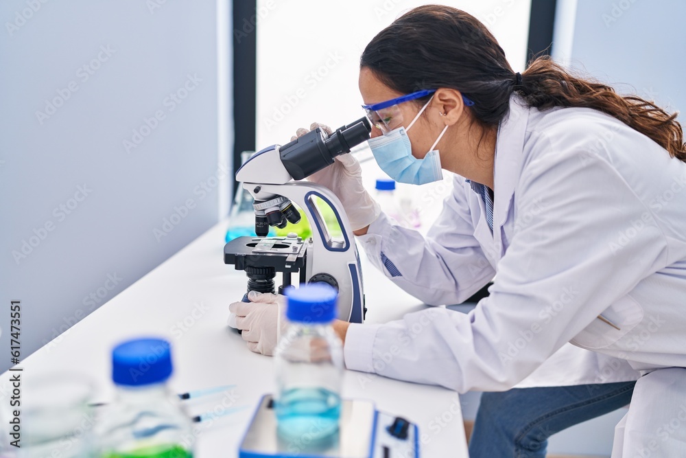 Wall mural young hispanic woman wearing scientist uniform using microscope at laboratory