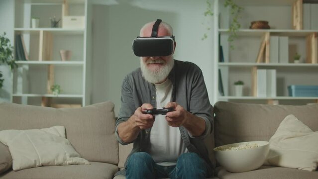 An Old Man in VR Glasses Plays Computer Games With a Joystick, Sitting in the Sofa and Eating a Popcorn.A Modern Elderly Man Plays PlayStation. Augmented Reality and Gaming for Seniors.