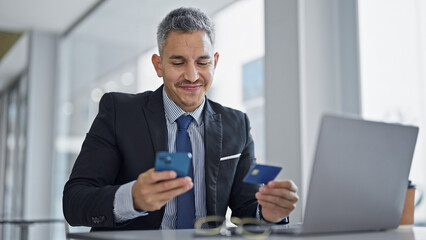 Fototapeta premium Young hispanic man business worker using smartphone holding credit card at office