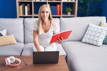 Young blonde woman student sitting on sofa reading book at home