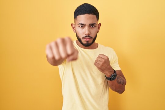 Young hispanic man standing over yellow background punching fist to fight, aggressive and angry attack, threat and violence