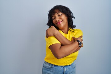 Hispanic woman standing over blue background hugging oneself happy and positive, smiling confident. self love and self care