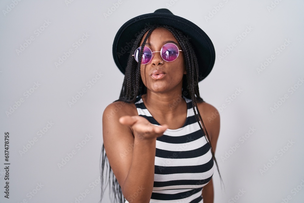 Poster young african american with braids wearing hat and sunglasses looking at the camera blowing a kiss w