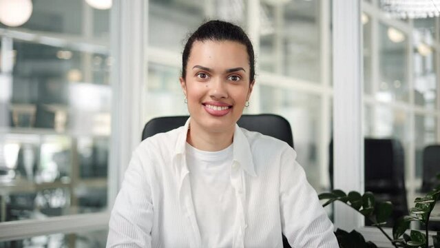 Smiling beautiful successful Latin American girl is sitting in office, looking into camera for a business portrait. Businesswoman with brown eyes, wearing a white shirt, posing for camera indoor.