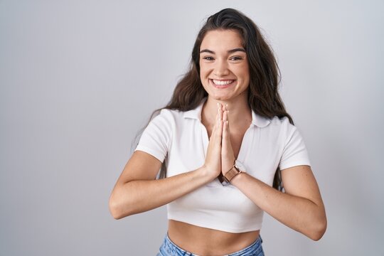 Young teenager girl standing over white background praying with hands together asking for forgiveness smiling confident.