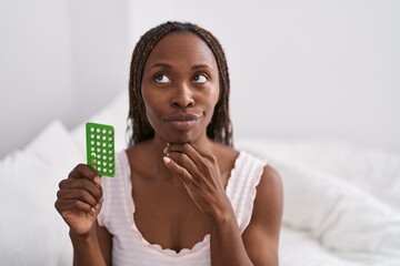 African american woman holding birth control pills serious face thinking about question with hand on chin, thoughtful about confusing idea