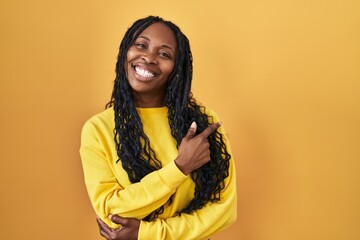 African woman standing over yellow background with a big smile on face, pointing with hand and finger to the side looking at the camera.