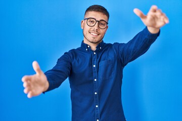 Naklejka premium Young hispanic man wearing glasses over blue background looking at the camera smiling with open arms for hug. cheerful expression embracing happiness.