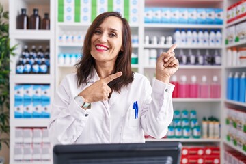 Middle age brunette woman working at pharmacy drugstore smiling and looking at the camera pointing with two hands and fingers to the side.