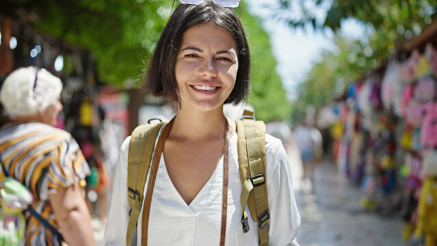 Young beautiful hispanic woman tourist smiling confident wearing backpack at street market