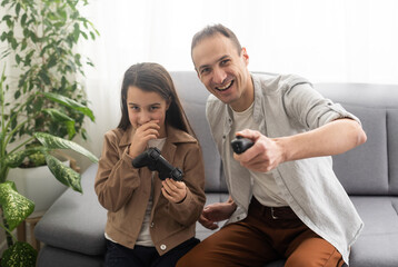 A father and a little daughter play video games using gamepads at home. A man and a child hold game joysticks in their hands. The family is having fun in the bedroom while being quarantined. © Angelov