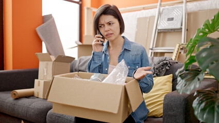 Young caucasian woman unpacking cardboard box speaking on the phone complaining at new home - obrazy, fototapety, plakaty