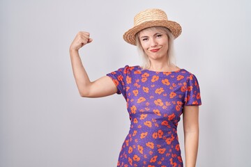 Young caucasian woman wearing flowers dress and summer hat strong person showing arm muscle,...