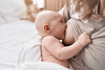 Adorable caucasian baby sitting on bed with mother sucking breast to eat at bedroom
