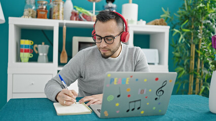 Hispanic man wearing headphones using laptop writing on notebook at dinning room