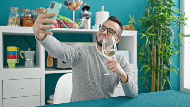 Hispanic man drinking glass of wine sitting on table taking selfie picture at dinning room
