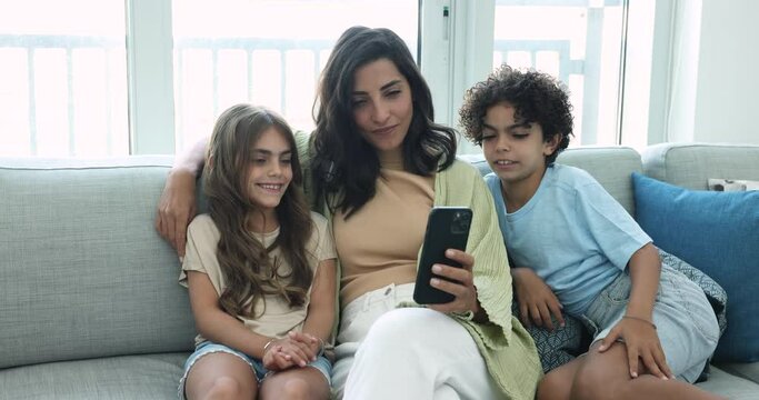 Young beautiful woman spend time with children sitting on cozy couch, making selfie photos on smartphone, use new filter or mobile application, watch funny video. Family pastime use modern technology