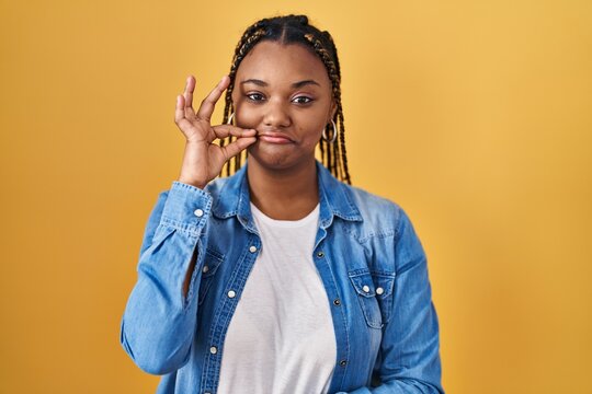African american woman with braids standing over yellow background mouth and lips shut as zip with fingers. secret and silent, taboo talking