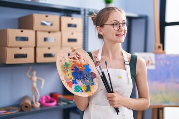 Young caucasian woman artist smiling confident holding paintbrushes and palette at art studio