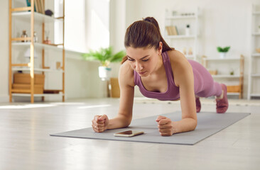 Beautiful fit young woman having sports workout at home, doing plank exercise on yoga mat, trying to endure physical exertion and using modern fitness mobile app on her cellular phone