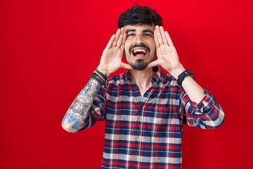 Young hispanic man with beard standing over red background smiling cheerful playing peek a boo with...