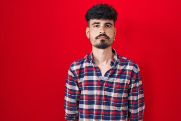 Obraz premium Young hispanic man with beard standing over red background relaxed with serious expression on face. simple and natural looking at the camera.