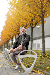 Portrait of a contented older gentleman amidst autumn trees. Mid-Adult stylish white bearded man, in a leather jacket, sitting on a bench and enjoying autumn's delight.