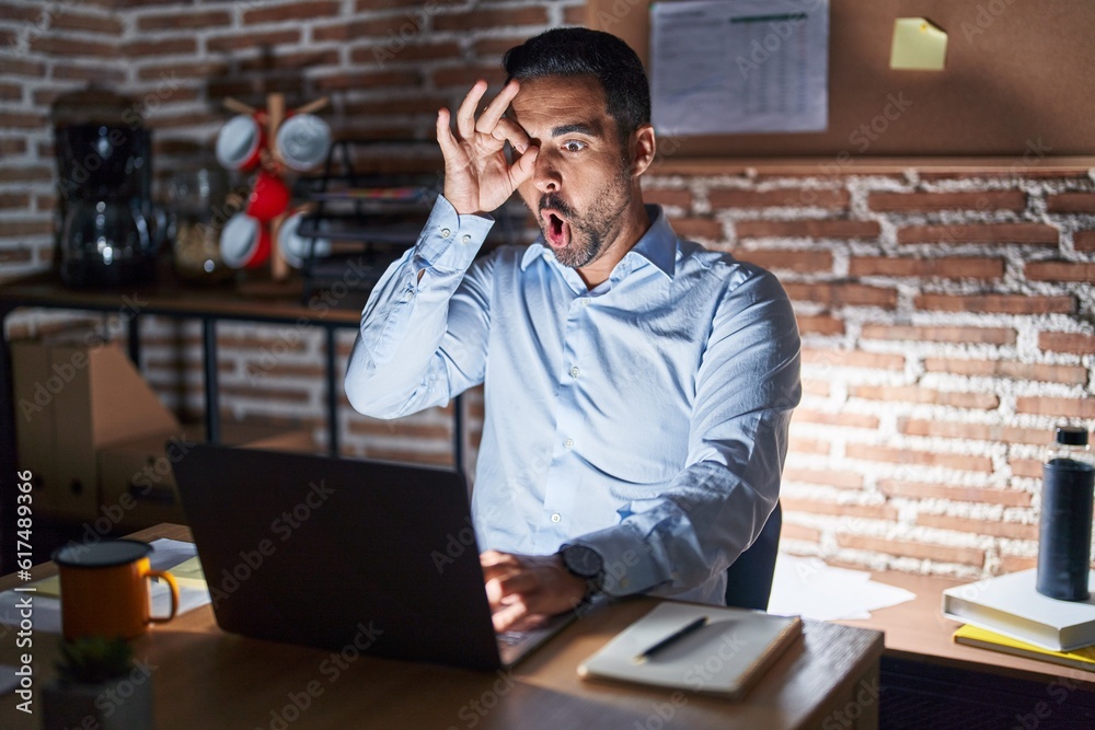 Poster Hispanic man with beard working at the office at night doing ok gesture shocked with surprised face, eye looking through fingers. unbelieving expression.