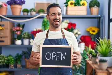 Hispanic man with beard working at florist holding open sign winking looking at the camera with...
