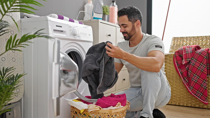 Young hispanic man smiling confident washing clothes at laundry room - obrazy, fototapety, plakaty
