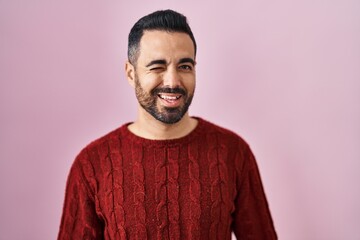 Young hispanic man with beard wearing casual sweater over pink background winking looking at the...