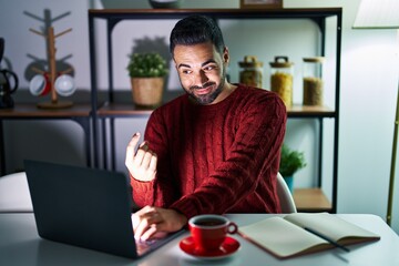 Young hispanic man with beard using computer laptop at night at home beckoning come here gesture with hand inviting welcoming happy and smiling