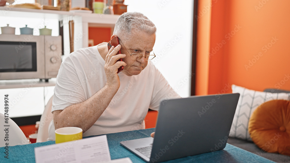 Sticker middle age grey-haired man talking on smartphone using laptop at dinning room