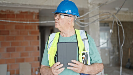 Middle age grey-haired man builder holding touchpad with relaxed expression at construction site