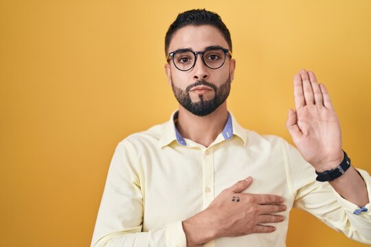 Hispanic young man wearing business clothes and glasses swearing with hand on chest and open palm, making a loyalty promise oath