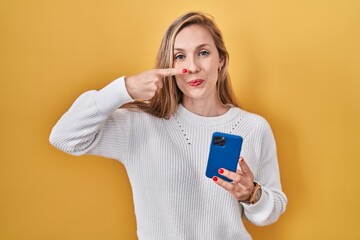 Young blonde woman using smartphone typing message pointing with hand finger to face and nose, smiling cheerful. beauty concept