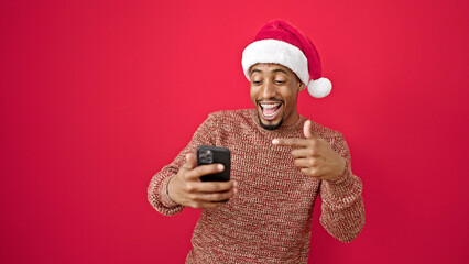 African american man wearing christmas hat pointing to smartphone surprised over isolated red background