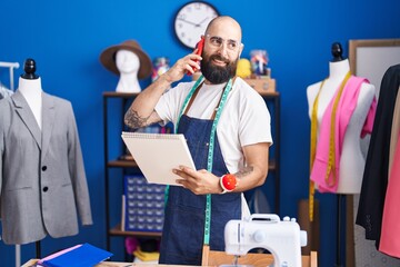 Young bald man tailor talking on smartphone looking clothing design at clothing factory