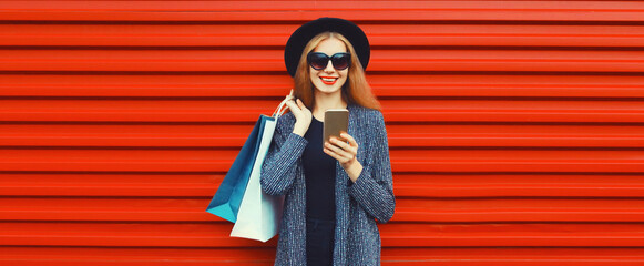Fototapeta premium Portrait of happy smiling woman with shopping bags and smartphone wearing blazer, black round hat on red background, blank copy space for advertising text