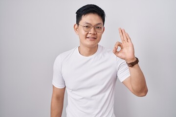 Young asian man standing over white background smiling positive doing ok sign with hand and fingers. successful expression.