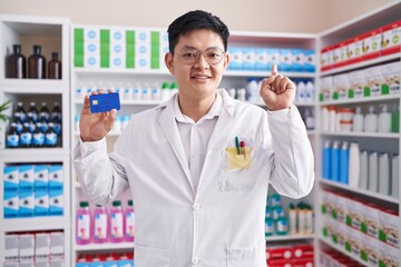 Young asian man working at pharmacy drugstore holding credit card surprised with an idea or question pointing finger with happy face, number one