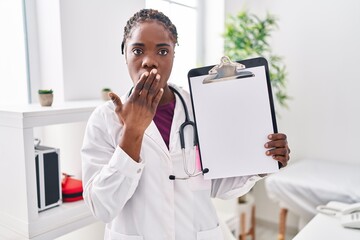 Beautiful black doctor woman holding clipboard covering mouth with hand, shocked and afraid for...
