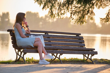 Lonely young woman sitting alone on lake shore bench enjoying warm summer evening. Wellbeing and relaxing in nature concept