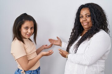 Mother and young daughter standing over white background inviting to enter smiling natural with open hand